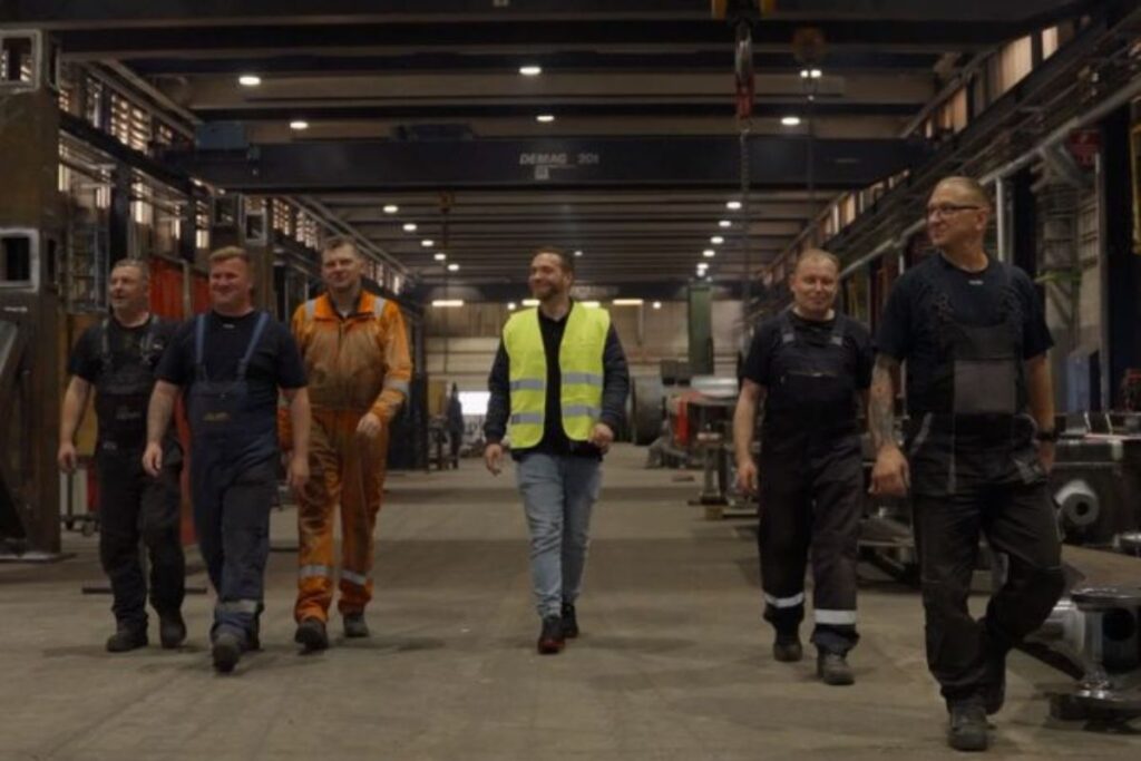 Gruppe af industriarbejdere og en koordinator i sikkerhedsvest, der går gennem en produktionshal – eksempel på bemanding og rekruttering i metal- og industribranchen.