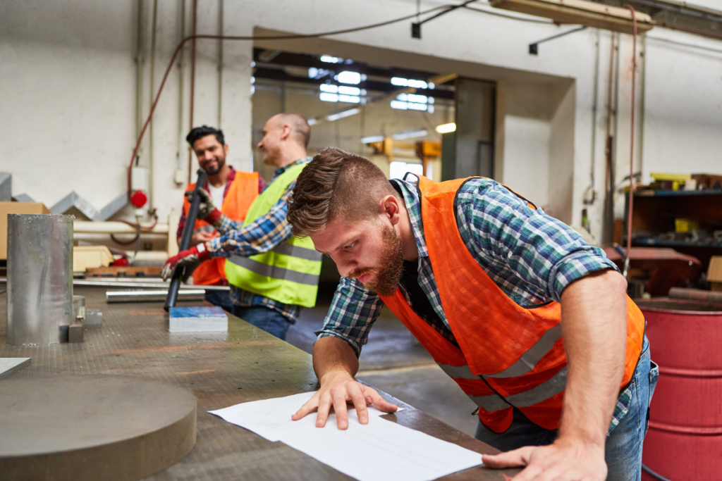Faglært industriarbejder gennemgår teknisk tegning i værkstedet, mens kolleger samarbejder i baggrunden.