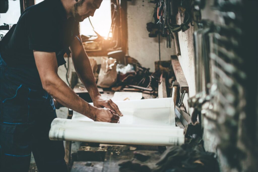 Håndværker arbejder ved arbejdsbord med tegninger i et værkstedsmiljø.