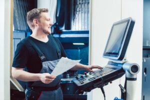 CNC-medarbejder ved styrepanel og teknisk tegning foran CNC-maskine i industriproduktion.